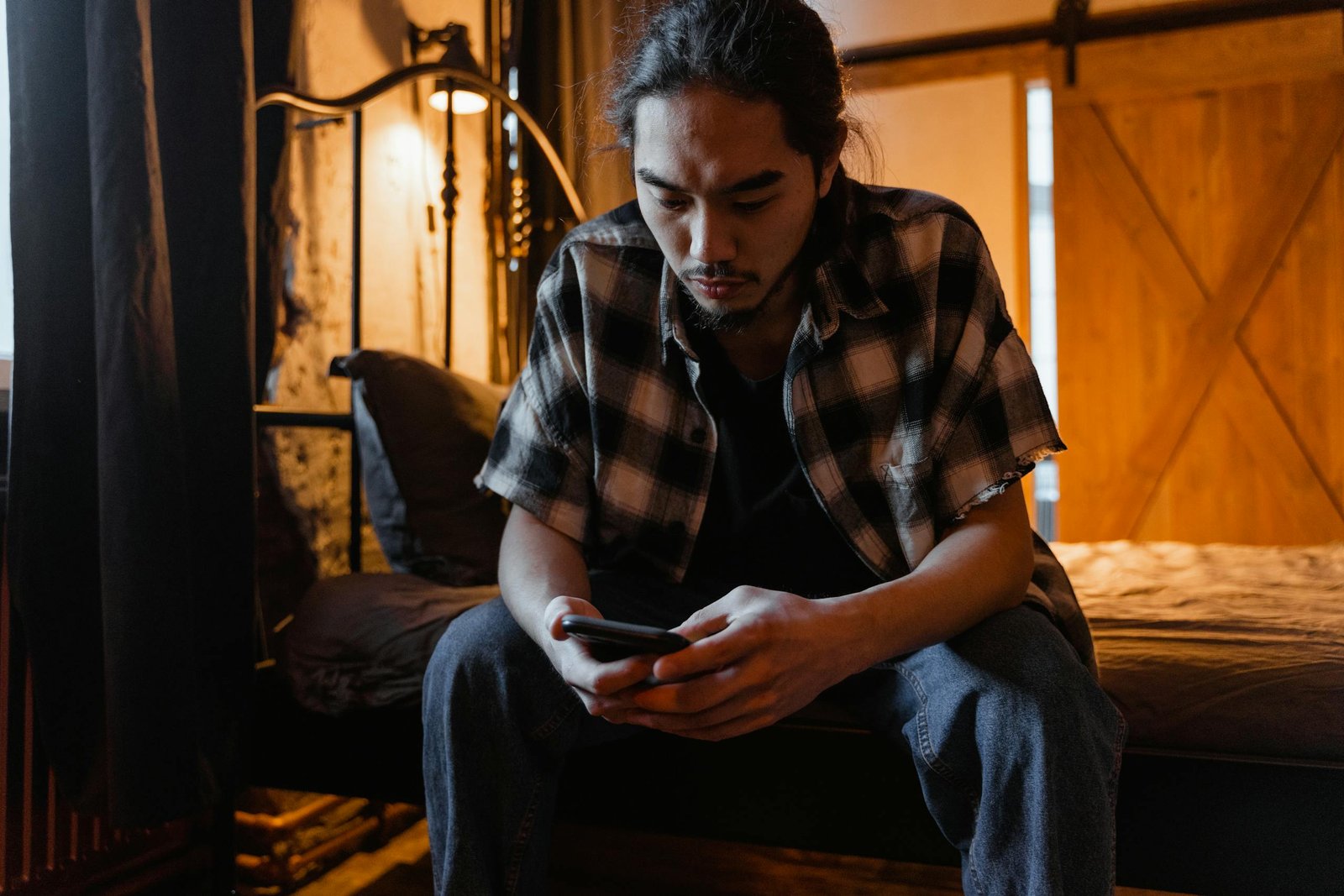 Pensive young man in casual attire using smartphone in dimly lit room, seated on bed.