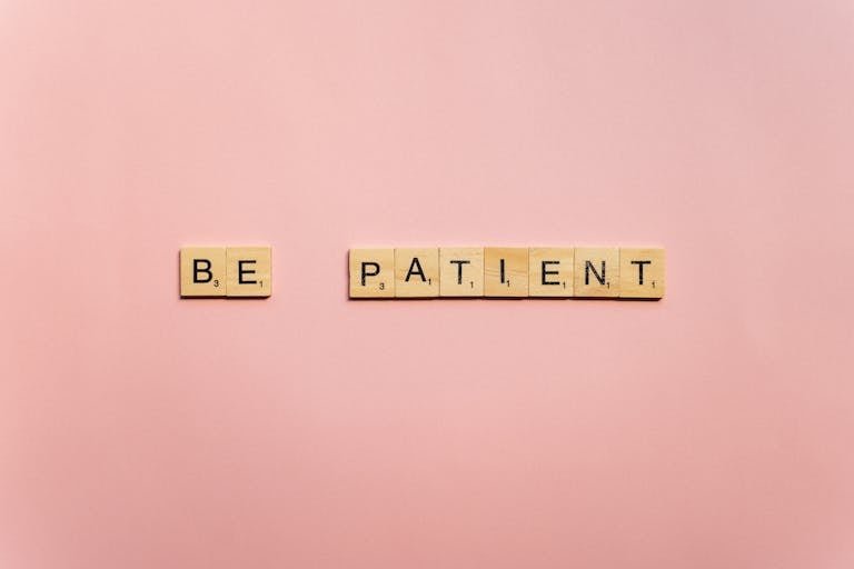 Inspirational words 'Be Patient' spelled with wooden tiles on a soft pink background.