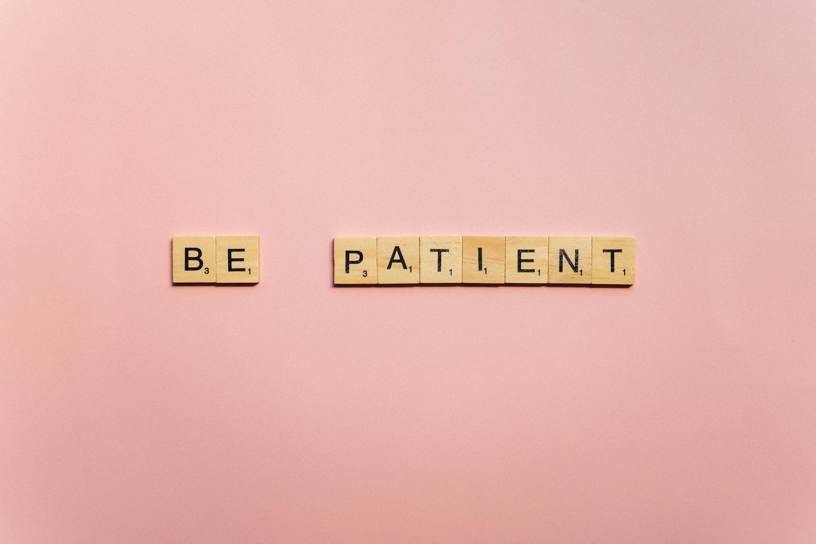 Inspirational words 'Be Patient' spelled with wooden tiles on a soft pink background.